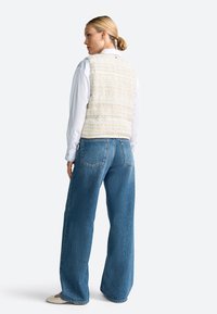 Gilet di tessuto crema chiaro sopra una camicia bianca, abbinato a jeans blu in denim larghi. Il gilet presenta un motivo a righe e un dettaglio con bottone sul retro.