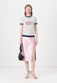 Camiseta de rayas con ribete azul marino y branding rojo, combinada con una falda midi rosa brillante que presenta una abertura lateral. Bolso negro y tacones beige.