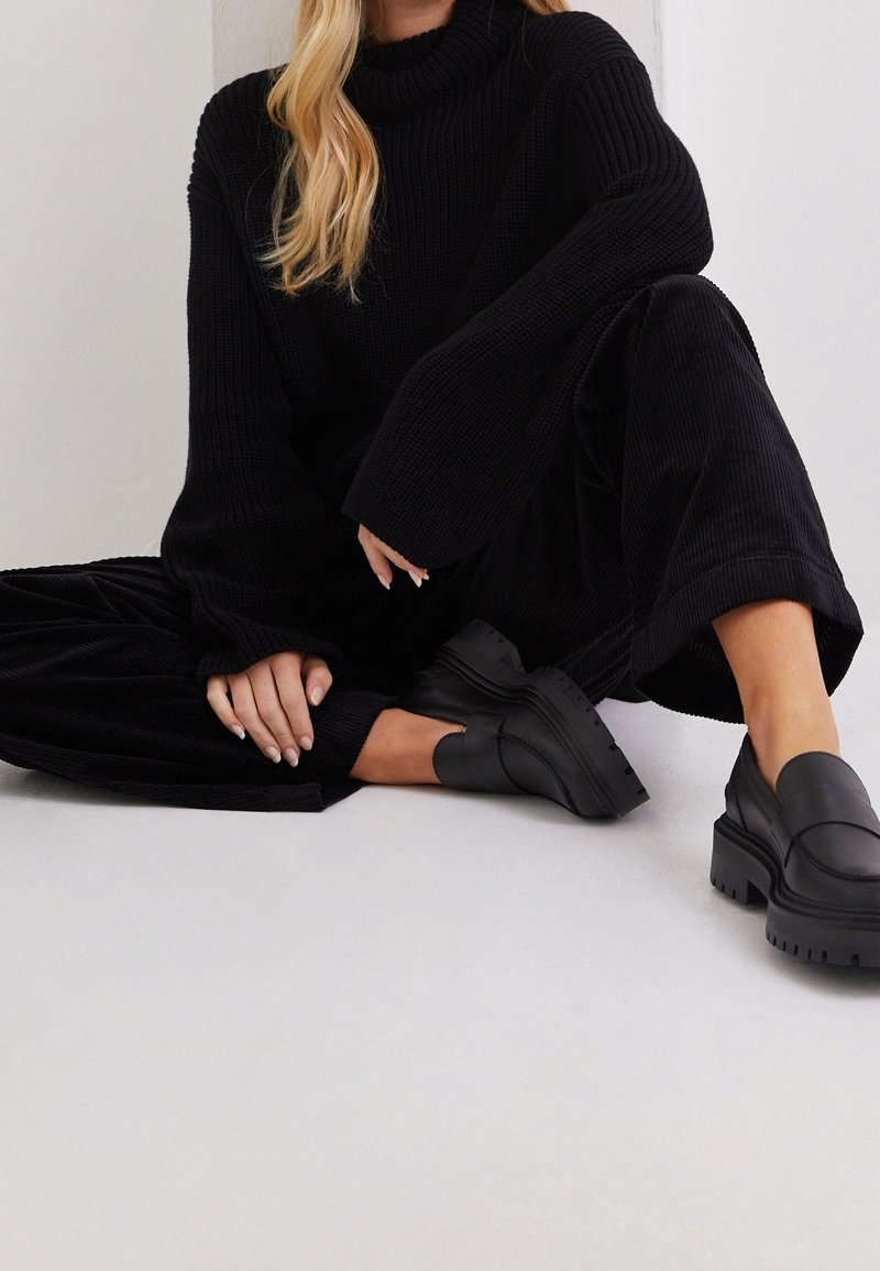 Pull en tricot côtelé noir avec un col roulé haut, associé à un pantalon en velours côtelé noir. Chaussures noires à enfiler épaisses avec des semelles lourdes.