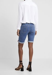 Mujer con shorts vaqueros azules remangados, blusa blanca de manga larga y sandalias negras de tacón grueso, de pie contra un fondo liso.