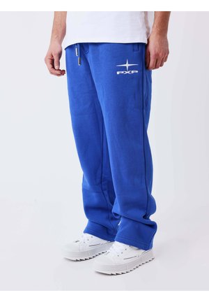 Uomo con pantaloni della tuta blu brillante con logo PXP e scarpe da ginnastica bianche, in piedi su uno sfondo chiaro e uniforme.