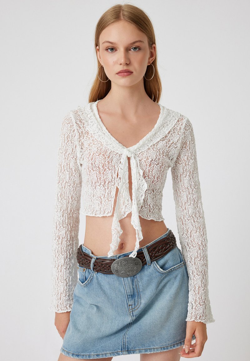 Valkoinen pitsinen crop-top, jossa on pitkät hihat, V-pääntie ja nauhakoteloedusta. Yhdistettynä farkkuiseen minihelmaan ja letitettyyn ruskeaan vyöhön.