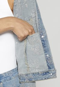 Vaaleansininen farkkutakki, jossa on kukkakirjontaa, erottuvaa ommelta ja pieni tasku sisähihassa. Napit näkyvät reunassa.