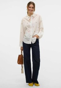 Camicia bianca con motivi a pois rossi, abbinata a jeans larghi blu scuro, scarpe gialle e una borsa marrone con tracolla.