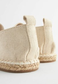 Beige canvas espadrilles with a textured surface, natural jute sole, and woven detailing at the base. Features small fabric heel loops.