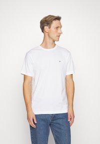 Mann trägt ein schlichtes weißes T-Shirt mit kurzen Ärmeln und blaue Jeans, steht vor einem weißen Hintergrund und schaut nach rechts.