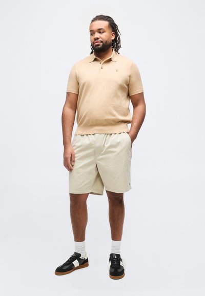 Άνδρας με dreadlocks στέκεται φορώντας μπεζ κοντομάνικο πόλο, ανοιχτόχρωμα χακί σορτς, άσπρες κάλτσες και μαύρα αθλητικά παπούτσια με άσπρες ρίγες.