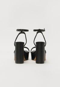 Sandales à talons hauts noirs avec des talons blocs épais, des brides de cheville et des orteils ouverts, présentant une finition en cuir lisse et un design minimaliste.