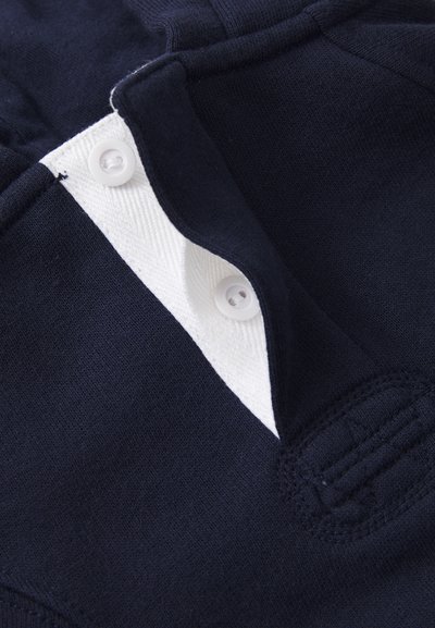 Lähikuva laivastonsinisestä kankaasta, jossa valkoinen nappilista ja kaksi valkoista muovinappia, näyttäen yksityiskohtaiset ompeleet ja tekstuurin.