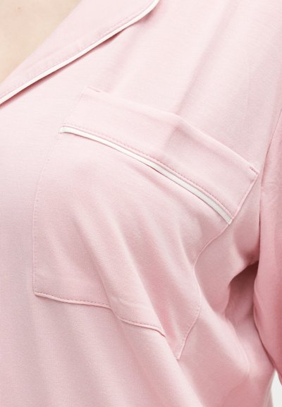 Κοντινή λήψη απαλού ροζ πουκαμίσου από ύφασμα με τσέπη στο στήθος περιγραμμένη με λευκό διακοσμητικό τελείωμα.
