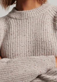 Pull en maille beige clair avec une texture côtelée, doté d'un col montant et de manches longues avec un motif tacheté subtil.