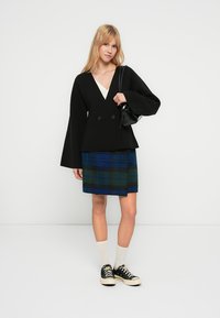 Black double-breasted jacket with wide sleeves, paired with a blue and green plaid skirt. Model wears black sneakers and white ankle socks.