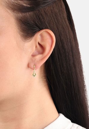 Donna con capelli scuri e lisci che indossa un piccolo orecchino a forma geometrica dorato con una pietra chiara centrale su uno sfondo neutro.