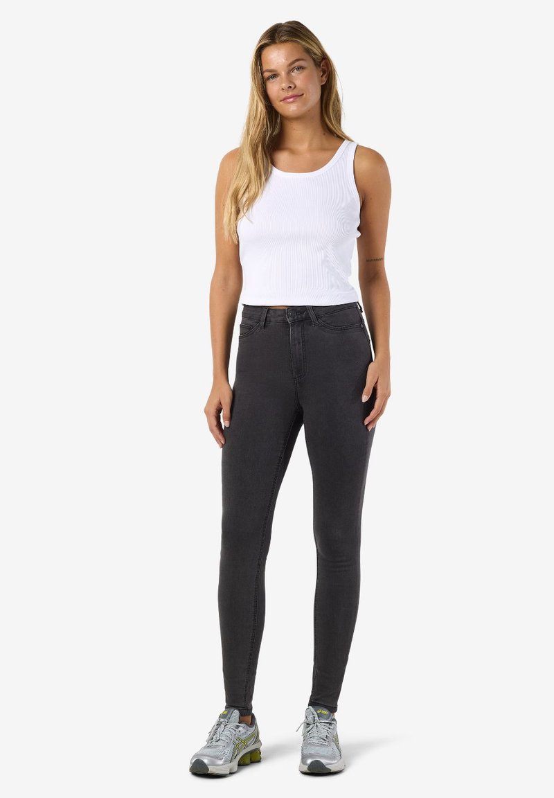 Juodos aukšto liemens siaurėjančios džinsai su lygiu paviršiumi, derinami su balta ribuota trikotažine palaidine be rankovių ir pilkais sportiniais bateliais.