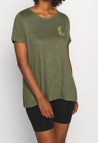 Olivenfarvet t-shirt med korte ærmer og løs pasform, pyntet med spredte perler og et struktureret dyreformet design på brystet.