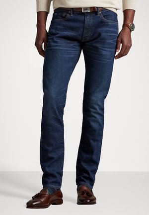 Mann trägt dunkelblaue, eng anliegende Jeans, braunen Ledergürtel, braune Tassel-Loafers und eine Armbanduhr am linken Handgelenk, steht vor einem einfarbigen Hintergrund.