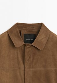 Veste en suède marron avec un col pointu et une étiquette noire visible portant la mention Massimo Dutti en blanc à l'intérieur de l'encolure.