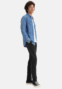 Camicia di jeans blu con bottoni a pressione, due tasche sul petto, indossata su una T-shirt bianca. Pantaloni neri e sneaker scure completano l'outfit.
