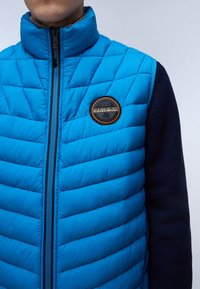 Blå vadderad väst med ett kviltat mönster, hög krage och svarta stickade ärmar. Har en cirkulär logotypapplikation på bröstet. Dragkedjestängning.