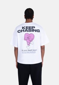 Hvit overdimensjonert t-skjorte med et stort rosa hjertegrafikk med hender, svart tekst "KEEP CHASING," og en humoristisk melding på baksiden.