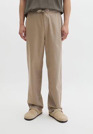 Mężczyzna ubrany w luźne beżowe spodnie ze sznurkiem i jasnobeżowe buty wsuwane, stojący na tle jednolitego jasnego tła.