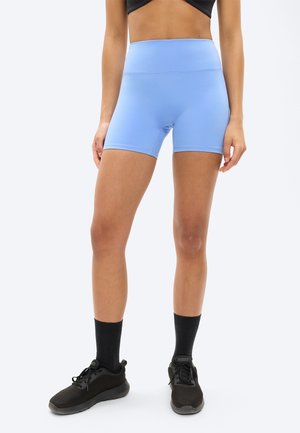 Žena oblečená v svetlomodrých športových šortkách s vysokým pásom, čiernych teniskách a čiernych ponožkách, stojaca na jednoduchom bielom pozadí.