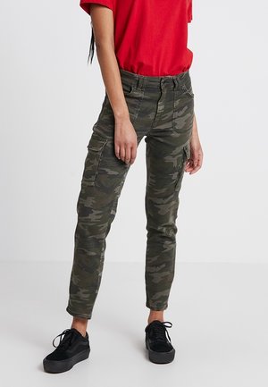 Person iført grønne camouflage cargobukser, rød kortærmet skjorte og sorte sneakers, stående mod hvid baggrund.