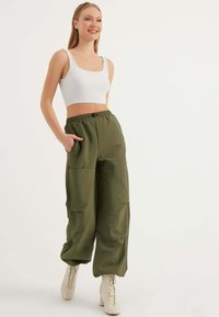 Olivengroene, wijde broek met een elastische tailleband en twee zijzakken, gecombineerd met een witte geribbelde crop top en lichtgekleurde enkellaarzen.