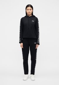 Γυναίκα που στέκεται φορώντας ένα μαύρο φούτερ EA7 Emporio Armani με κουκούλα και αντίστοιχο παντελόνι φόρμας με λευκά λογότυπα στα μανίκια και τις παντελόνια, και λευκά αθλητικά παπούτσια.