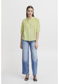 Blusa verde chiaro con bottoni, maniche corte a sbuffo e scollatura arricciata, abbinata a jeans in denim blu larghi e sandali neri con tacco.