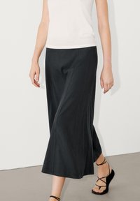 Leveled dark trousers yang lebar terbuat dari kain halus, dilengkapi dengan detail jahitan vertikal, dipadukan dengan sandal hitam bertali dan atasan putih.