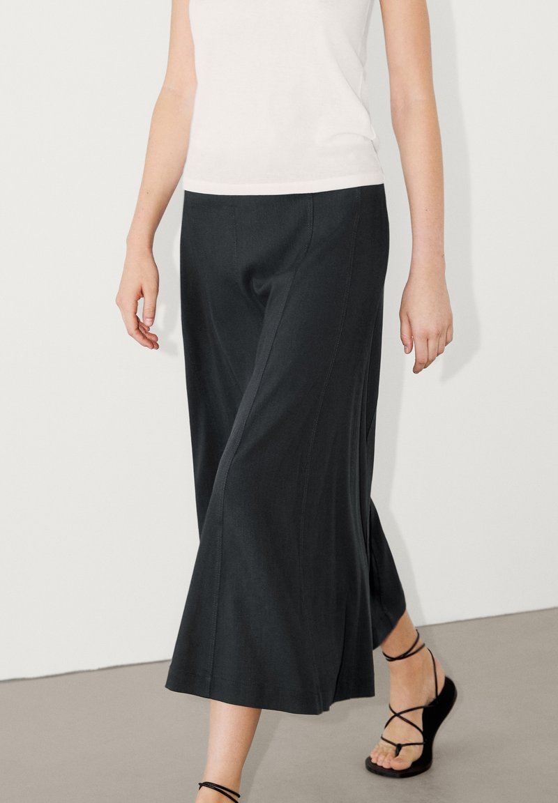 Leveled dark trousers yang lebar terbuat dari kain halus, dilengkapi dengan detail jahitan vertikal, dipadukan dengan sandal hitam bertali dan atasan putih.