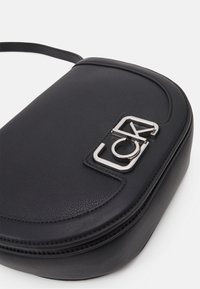 Czarna skórzana torba na ramię o zaokrąglonym kształcie, z zamkiem błyskawicznym i srebrnym logo na przodzie, z gładką fakturą.