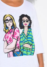 T-shirt bianco con stampa grafica raffigurante due donne dei cartoni animati. Una indossa un top floreale rosa, mentre l'altra indossa un top avvolgente a righe verdi.