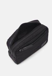 Trousse de toilette en nylon noir au design épuré, avec deux compartiments zippés, un accent en cuir texturé et une étiquette intérieure.