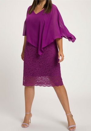 Kvinde iført en lilla lagdelt kjole med en gennemsigtig kappe-lignende top og blonder nederdel, kombineret med lavendel høje hæle sandaler.