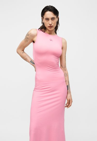 Γυναίκα με σκούρα, υγρή όψη μαλλιά, φοράει ροζ στενή αμάνικη φόρεμα, ποζάρει με το ένα χέρι στη μέση και έχει ορατά τατουάζ στα χέρια.