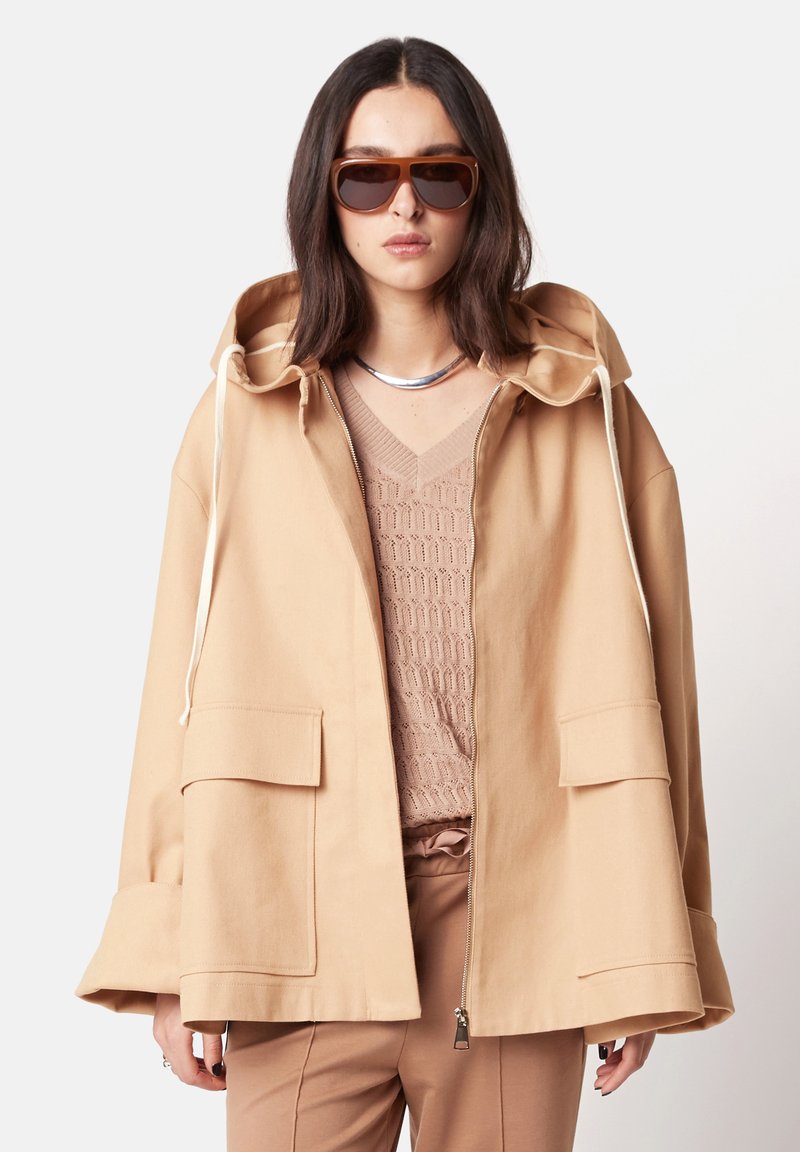 Hellbeige Jacke mit Kapuze und Zugband, zwei aufgesetzten Taschen vorne und Reißverschluss. Getragen über einem strukturierten beigen Oberteil. Braune Sonnenbrille rundet den Look ab.