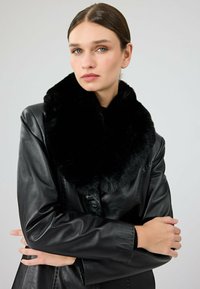 Czarna skórzana kurtka z szerokim, czarnym kołnierzem z sztucznego futra. Posiada zapięcie na guziki oraz miękką, gładką teksturę. Modelka pozuje z założonymi rękami.