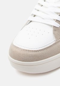 Κοντινό πλάνο μύτης αθλητικού παπουτσιού από μπεζ σουέτ και λευκό δέρμα με λευκά κορδόνια και λεπτομέρειες διάτρησης στο δέρμα.