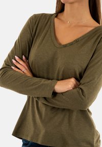 Camicia a maniche lunghe verde oliva con scollo a V, dotata di tre bottoni sul collo e una vestibilità comoda. Texture morbida e dettagli di orlo senza cuciture.