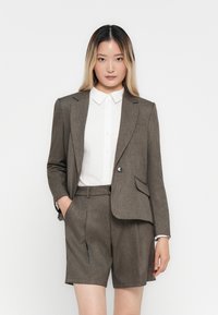 Jonge vrouw in een bruine blazer en bijpassende shorts, met een wit overhemd, staand met één hand in haar zak tegen een witte achtergrond.