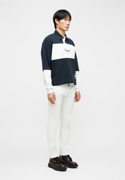 Νεαρός άνδρας που στέκεται πλευρικά, φοράει μια μακρυμάνικη μαύρη και λευκή μπλούζα, λευκό παντελόνι και σκούρα καφέ δερμάτινα παπούτσια με κορδόνια σε απλό φόντο.