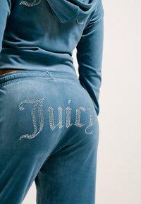 Zila velūra treniņtērps, kas ietver kapuci ar akmens rotājumiem un atbilstošas bikses. Vārds "Juicy" ir izveidots ar akmeņiem uz muguras.