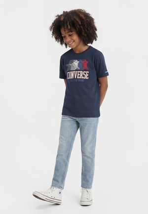 Enfant aux cheveux bouclés souriant, portant un t-shirt Converse bleu marine avec des motifs de baskets, un jean bleu clair et des baskets montantes blanches.