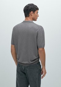Polo grigia con una texture morbida, maniche corte e colletto classico. Indossata con jeans scuri, vista da dietro.
