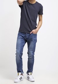 Marinblå kortärmad t-shirt med små vita prickar, kombinerad med blå jeans och vita sneakers med svarta ränder och logotyp.