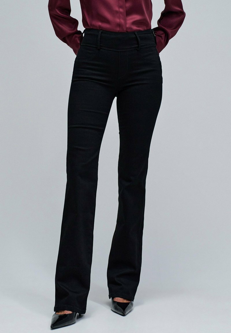 Pantalones acampanados negros hechos de una tela elástica, con una cintura alta y diseño sin costuras, combinados con tacones negros puntiagudos.