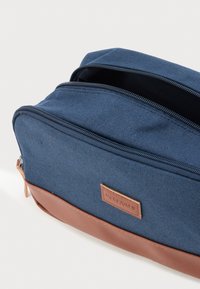 Μπλε τσάντα από καμβά με καφέ δερμάτινη βάση και κλείσιμο με φερμουάρ. Διαθέτει ετικέτα με λογότυπο και υφή ύφασματος.