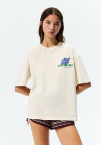 Cream-colored cotton t-shirt with short sleeves, featuring a blue shell graphic and green text that reads "CALM DEEP SEA."